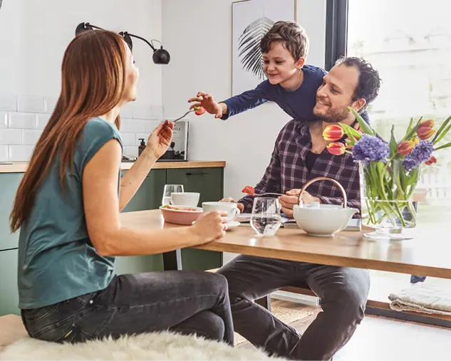 Familie sitzt gemeinsam am Holztisch in moderner Küche. Eltern und Kind genießen eine Mahlzeit in heller Wohnküche mit natürlichem Licht, Blumen und wohnlicher Atmosphäre. Küche als Mittelpunkt des Familienlebens.